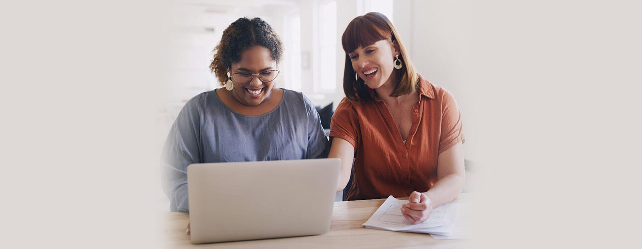 Zwei Frauen sitzen vor lächelnd vor einem Notebook. Eine Frau zeigt der anderen etwas am Bildschirm.