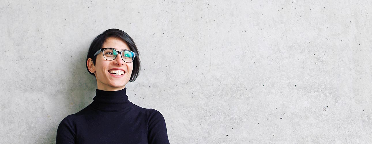 Frauenberufszentrum*Vorarlberg Eine Frau mit schwarzem Pagenschnitt und Brille lehnt an einer grauen Wand und blickt lächelnd in die Ferne.