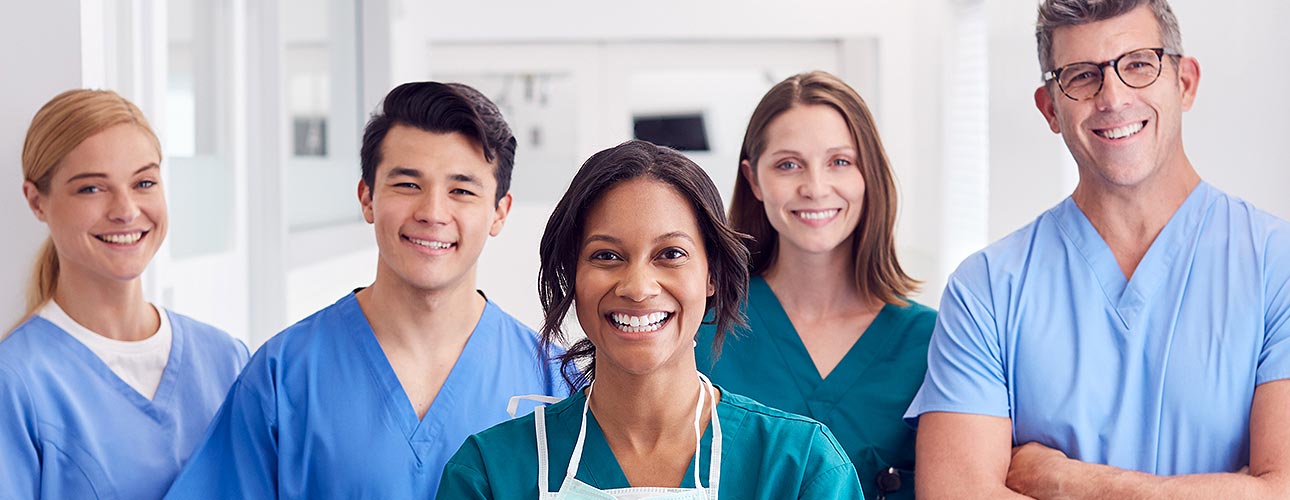 Ein Pfleger*innen- und Ärzt*innen-Team, bestehend aus drei Frauen und zwei Männern, lächelt in die Kamera. Alle tragen ihre Pflege-Uniform. Im Hintergrund sieht man einen weißen Gang mit einer Türe.