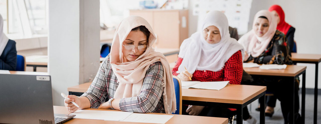 4 Frauen sitzen schreibend hintereinander an Tischen in einem Kurs. Sie tragen bunte Kopftücher.