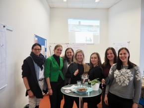 Eine Gruppe von Frauen lächelt freundlich in die Kamera. Vor ihnen steht ein Glastisch mit zwei Gläsern und eine Wasserkanne.