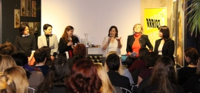 Fünf Frauen sitzen vor einem Publikum. Eine davon hält ein Mikrofon und spricht zum Publikum. 