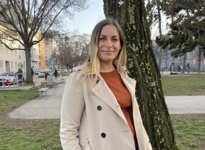Alexandra Stolz hat das das Kolleg für Wirtschaftsingenieurwesen am TGM abgeschlossen. In ihrer Arbeit kombiniert sie Technik und Wirtschaft. Absolventin des Fit-Programms steht in einem Mantel vor einem Baum und lächelt in die Kamera.