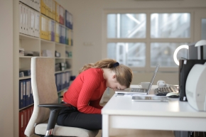 Eine Frau sitzt an einem Schreibtisch in einem Büro und hat ihren Kopf erschöpft auf den Laptop gelegt.