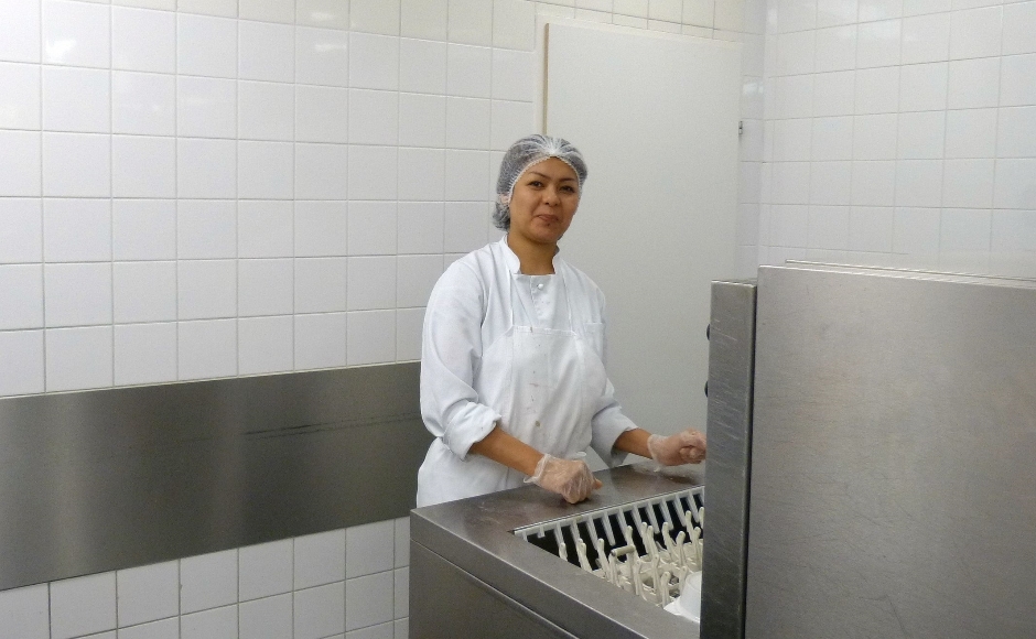 Eine Frau mit einem Haarnetz auf dem Kopf lächelt in die Kamera. Sie steht neben einem Gastro-Geschirrspüler.