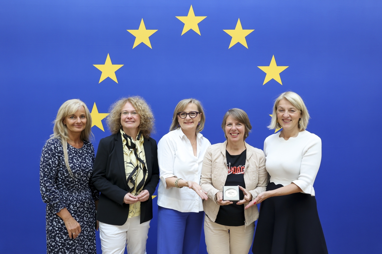 Fünf Frauen lächeln vor einer EU-Flagge. Die mittlere Frau hält eine Medaille in einer Box, die ihr von den anderen präsentiert wird. Alle sind elegant gekleidet, die Stimmung ist feierlich.