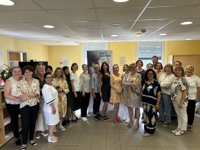Eine Gruppe von 20 Frauen und einem Mann steht dicht beisammen in einem hellen Raum, alle lächeln freundlich. Im Hintergrund ist ein Poster mit der Aufschrift "ABZ Austria" sichtbar. Die Stimmung wirkt fröhlich und harmonisch.