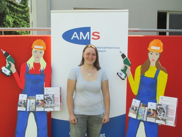 Eine junge Frau steht lächelnd vor einem AMS-Aufsteller. Links und rechts von ihr stehen Papp-Aufsteller, die Handwerkerinnen zeigen.